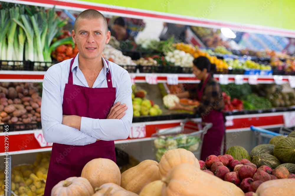 Φωτογραφία Supermarker worker in apron standing in salesroom among shelves with fruits and vegetables and looking in camera