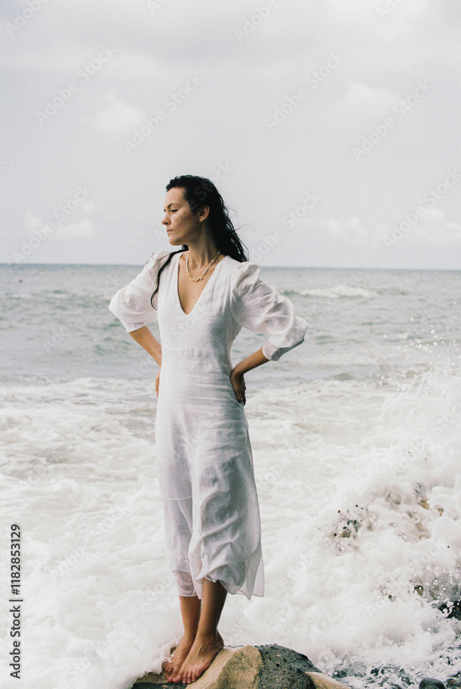 A young woman wearing a white dress, standing on a rock in the sea A young woman wearing a white dress, standing on a rock in the sea