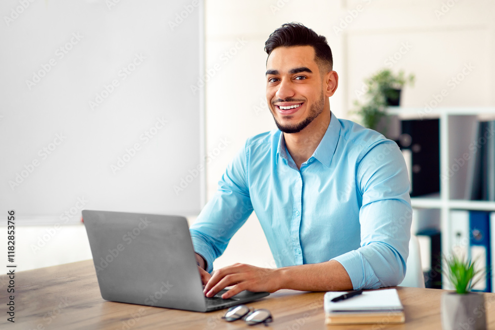 Fototapeta premium Attractive young Arab businessman using laptop pc at his desk in home office. Cheerful Eastern guy entrepreneur typing on computer keyboard, having in online conference at workplace