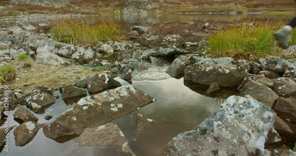 Hiker in a vibrant yellow jacket jumping over rocky terrain and puddles, reflecting the sky in a stunning mountain landscape featuring a serene lake and lush grass Hiker in a vibrant yellow jacket jumping over rocky terrain and puddles, reflecting the sky in a stunning mountain landscape featuring a serene lake and lush grass