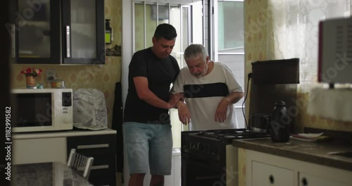 A man supporting an elderly man with dementia or age-related illness walking forward in a kitchen, showing care, assistance, and compassion in a home environment