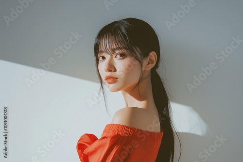 A young Asian woman with dark hair in a low ponytail, wearing an offshoulder orange dress, is captured in a sunlit portrait against a minimalist lightgrey background. The lighting creates soft shadows photo