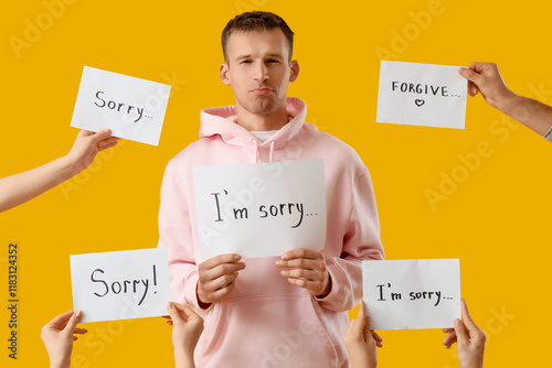 Sad young man and hands holding paper sheets with apologies on orange background photo