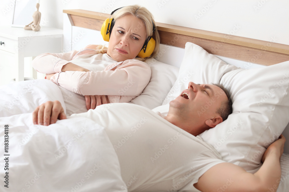 Irritated mature woman in hearing protectors lying with her snoring husband on bed at home Irritated mature woman in hearing protectors lying with her snoring husband on bed at home
