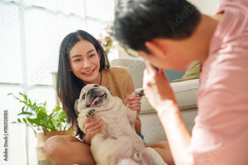 Asian woman smiles as she cuddles her cheerful pug dog while a man taking a photo with camera in living room.