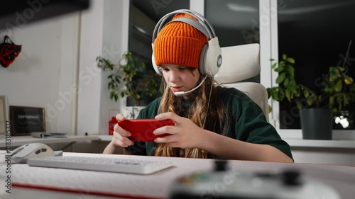 Focused female gamer in headphones immersed in video game session on smartphone. Gamingpro and leader, showcasing skills tournament setting. Perfect energy and style for gaming enthusiasts and teens
