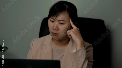 A woman in a light blazer sits at a desk, looking stressed while working on a laptop, with her hand on her temple.