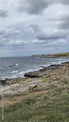 This serene coastal scene features a rugged shoreline, gentle waves, vibrant green grass, and a beautiful cloudy sky, creating a picturesque view for nature lovers to enjoy