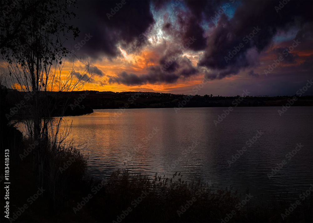 Fototapeta premium Stunning lakeside sunset with dramatic clouds, vibrant colors reflecting on the water, and silhouetted trees creating a peaceful evening scene