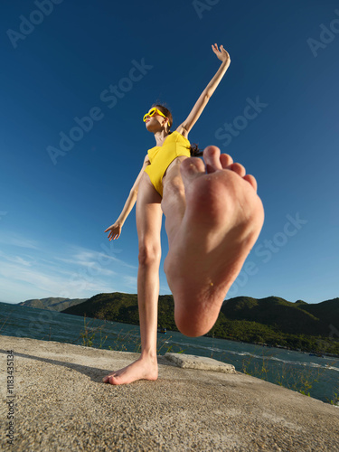 A woman in a yellow swimsuit performs a handstand on a concrete wall with a clear blue sky in the background, showcasing strength, balance, and outdoor fitness on a sunny day photo