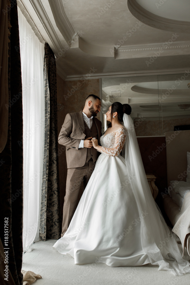 The bride and groom stand next to each other, holding hands and looking at each other with smiles. In the background you can see part of the room with large windows The bride and groom stand next to each other, holding hands and looking at each other with smiles. In the background you can see part of the room with large windows