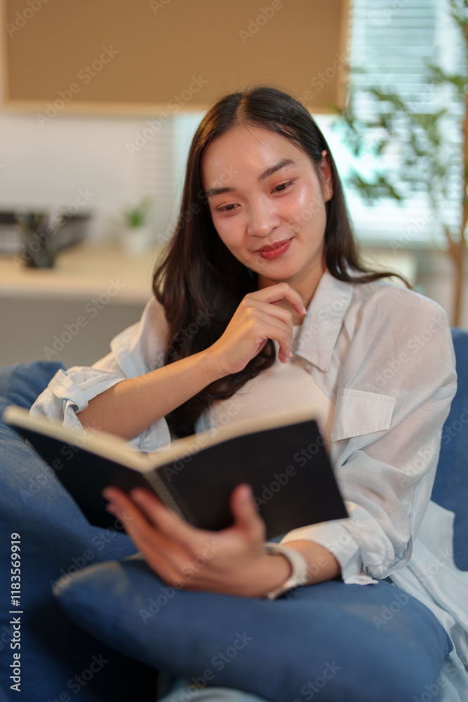 Fototapeta premium Comfortable young asian woman enjoying reading interesting book while sitting on sofa in cozy living room at home, spending leisure time with pleasure