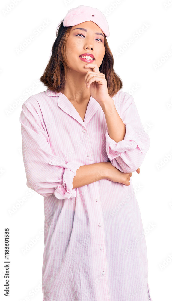 Young beautiful chinese girl wearing sleep mask and pajama looking confident at the camera with smile with crossed arms and hand raised on chin. thinking positive. Young beautiful chinese girl wearing sleep mask and pajama looking confident at the camera with smile with crossed arms and hand raised on chin. thinking positive.