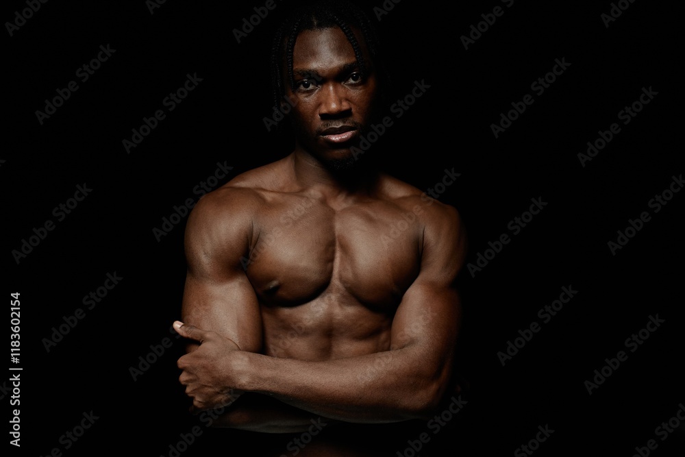 Fototapeta premium Medium portrait shot of shirtless African American man with muscular torso posing with arms crossed confidently looking at camera in dimly lit studio against black background, copy space