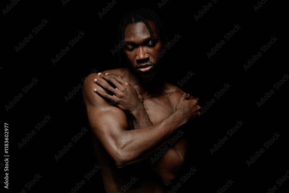 Medium portrait shot of Black man with eyes closed gently embracing bare shoulders in peaceful feeling of self acceptance while posing against black background in contrast light, copy space Medium portrait shot of Black man with eyes closed gently embracing bare shoulders in peaceful feeling of self acceptance while posing against black background in contrast light, copy space