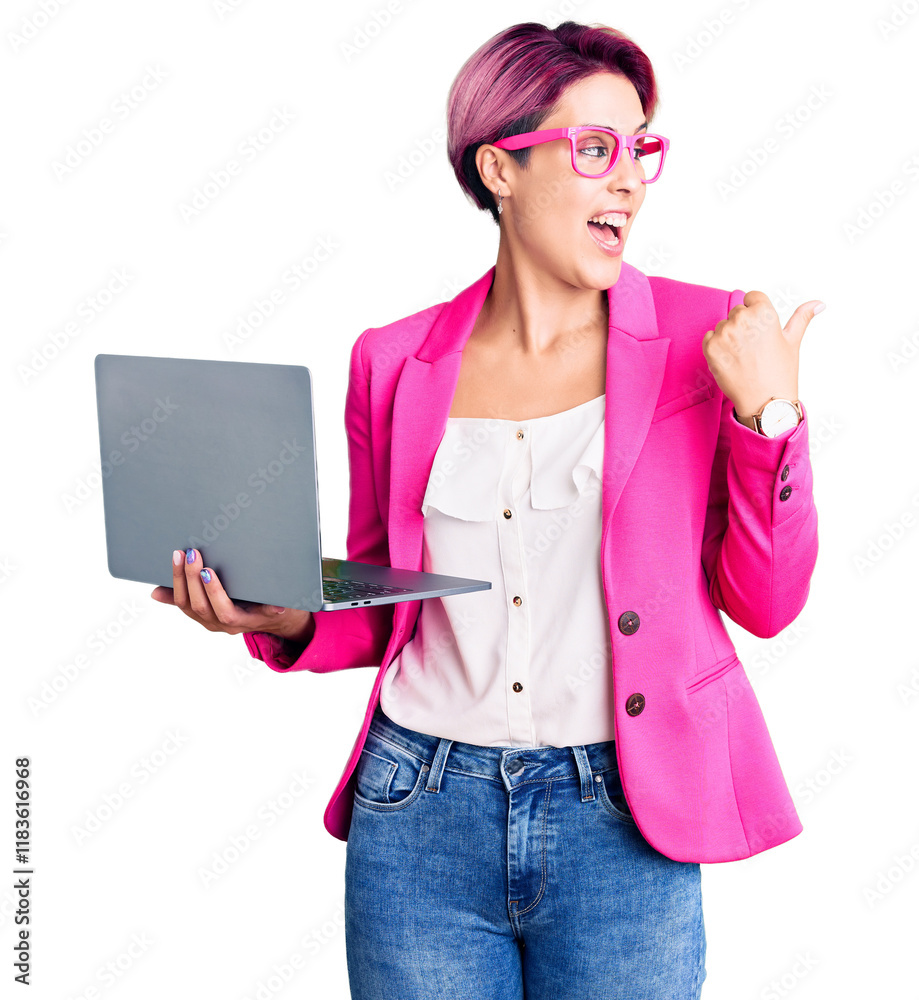 Young beautiful woman with pink hair wearing glasses holding laptop pointing thumb up to the side smiling happy with open mouth Young beautiful woman with pink hair wearing glasses holding laptop pointing thumb up to the side smiling happy with open mouth