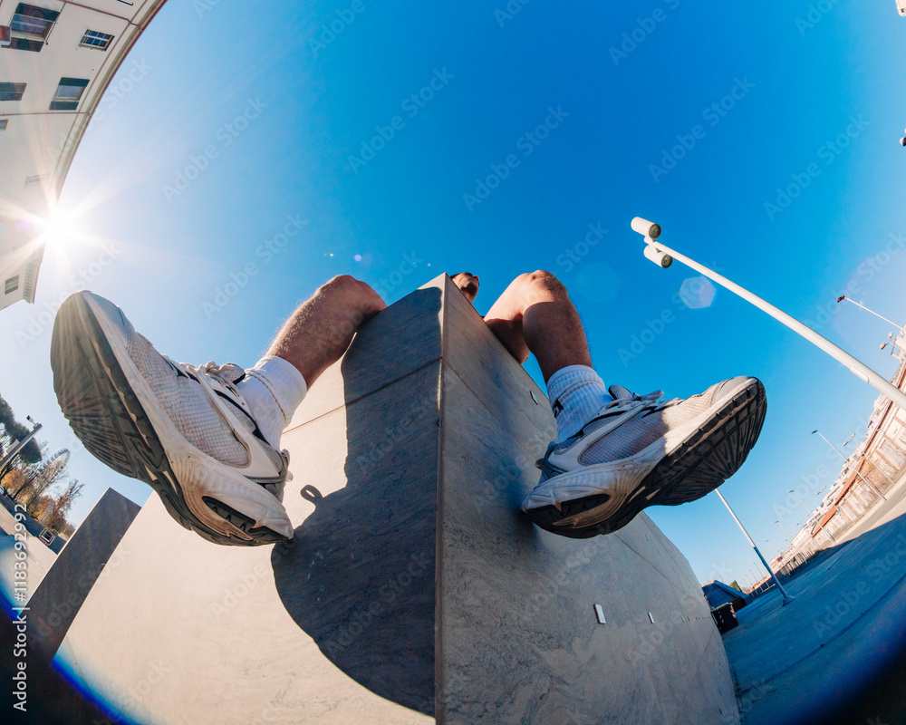 © Studio Marmellata - A fisheye lens close-up showing a young man's sneakers resting on a concrete ledge in an outdoor urban area with bright sunlight and clear blue skies in the background © Studio Marmellata - A fisheye lens close-up showing a young man's sneakers resting on a concrete ledge in an outdoor urban area with bright sunlight and clear blue skies in the background