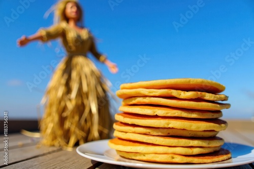 A stack of golden pancakes is placed beside a straw effigy of a woman. In the background, a bright blue sky enhances the festive atmosphere of the Maslenitsa celebration