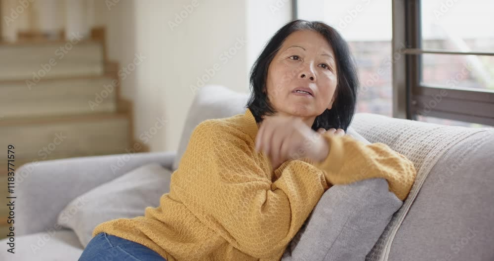 Senior Asian woman in yellow sweater sitting on couch, looking thoughtful at home Senior Asian woman in yellow sweater sitting on couch, looking thoughtful at home