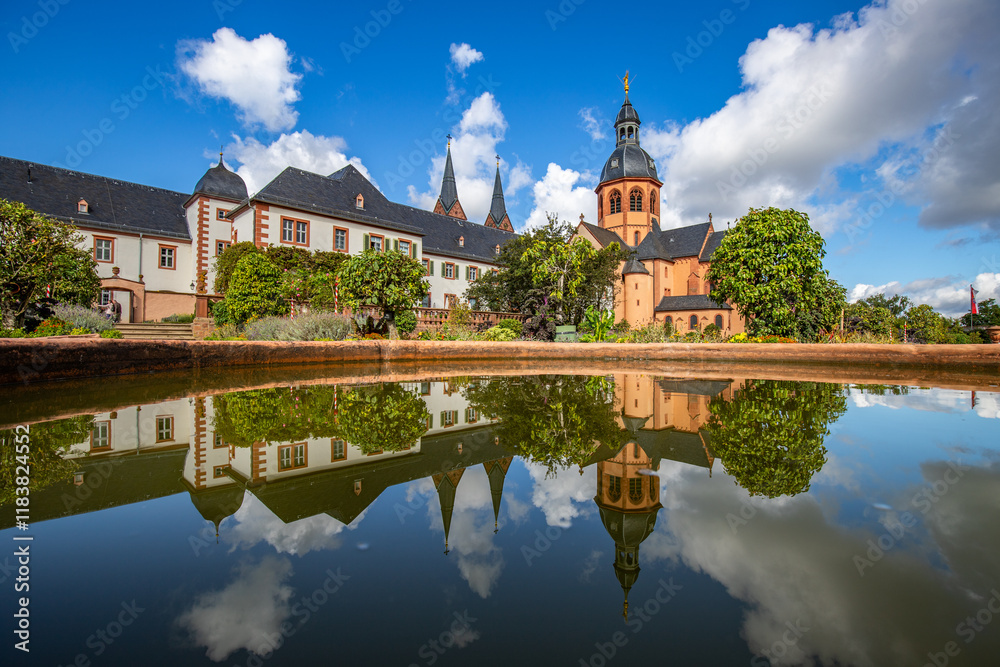 A historic monastery in the morning. Monastery walls, church and buildings are reflected in the water of a fountain. Beautiful garden in Seligenstadt Monastery, Germany A historic monastery in the morning. Monastery walls, church and buildings are reflected in the water of a fountain. Beautiful garden in Seligenstadt Monastery, Germany
