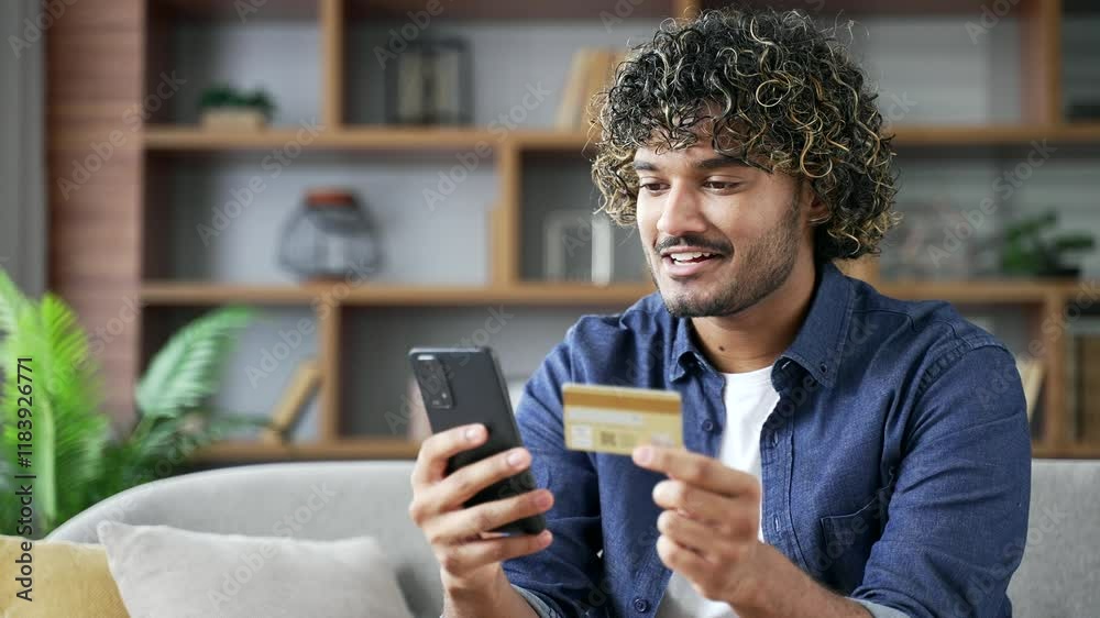 Happy young man doing online shopping typing credit card data on mobile phone sitting on sofa in living room at home. Smiling male makes a purchase, satisfied with a successful transaction. Close up