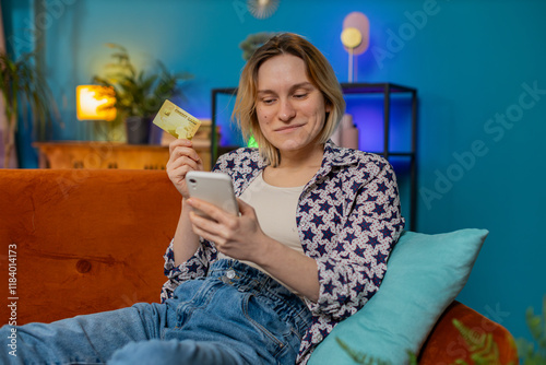 Happy young woman doing payments online purchase shopping in internet store. Smiling Caucasian girl holding banking credit card involved in verification process in mobile application on couch at home.