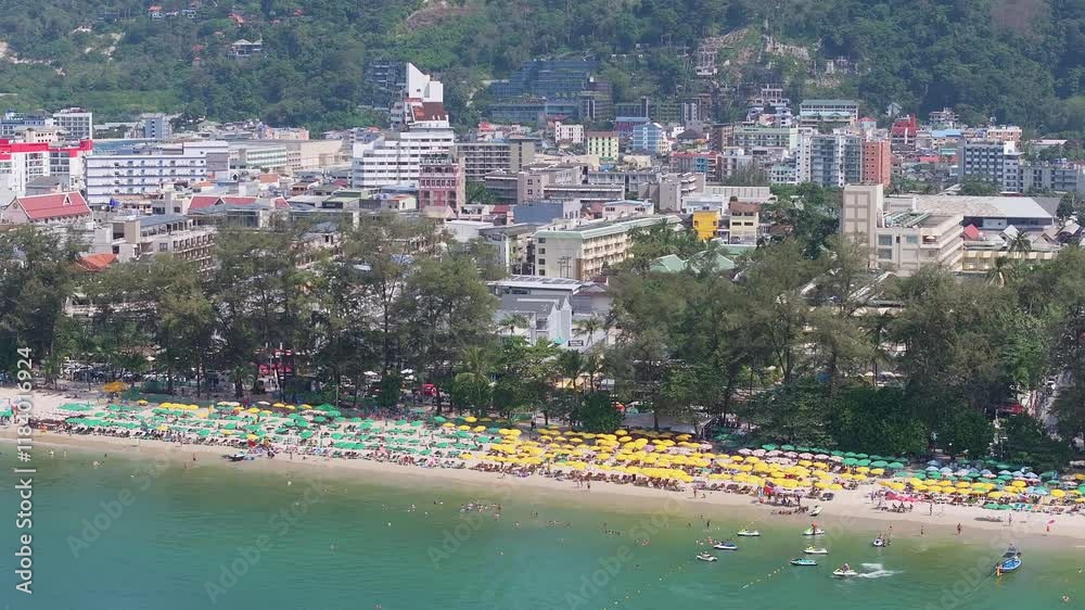 Aerial view of Patong Beach, Phuket, showing green and yellow umbrellas, visitors, turquoise waters, jet skis, and urban buildings by lush hills. Aerial view of Patong Beach, Phuket, showing green and yellow umbrellas, visitors, turquoise waters, jet skis, and urban buildings by lush hills.