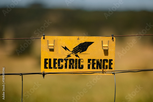 Metal fence with yellow 'Electric Fence' sign in rural landscape under clear blue sky photo