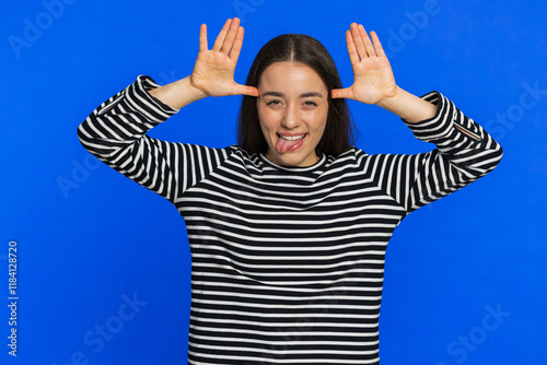 Funny Caucasian woman showing tongue making faces at camera, fooling around, joking, aping with silly face, teasing, bullying, abuse disrespect. Pretty young girl isolated on blue background indoors