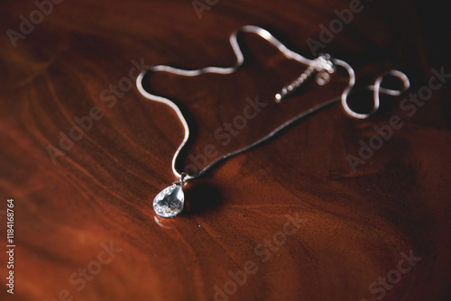 A silver necklace with a crystal pendant sits on a wooden table. The pendant is shaped like a teardrop and is surrounded by a chain. The necklace appears to be a piece of jewelry