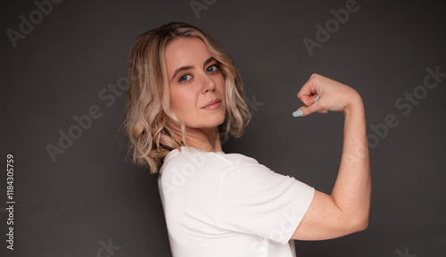 Confident woman flexing her arm in a strong pose, showcasing empowerment and determination. Blonde hair, casual white shirt, and a neutral background emphasize strength and self-confidence.