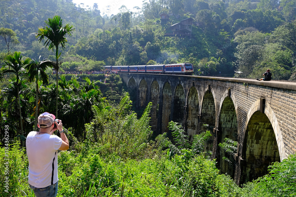 Man in white shirt taking photo of train passes iconic Nine Arch Bridge in Ella, Nuwara Eliya, Sri Lanka