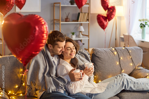 Valentine day, happy loving couple on sofa, man, woman holding hot drink, sharing tender love, light garland, heart shaped red balloons foil decor, wedding party room decoration, romantic gift  photo