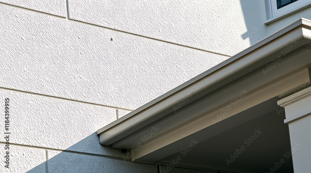Obraz premium Close-up view of a house exterior, focusing on a light beige rain gutter and fascia board against a textured, off-white siding. A portion of a covered porch and column are also visible.
