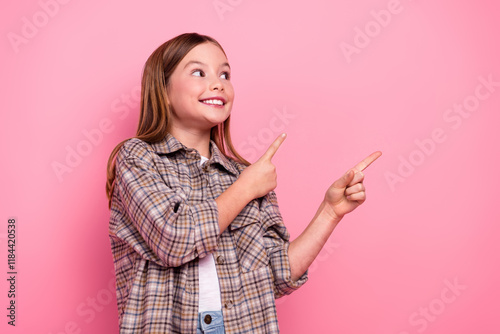 Young girl pointing sideways with a smile, dressed in a trendy checkered shirt against a pink background conveying happiness and lighthearted energy. photo