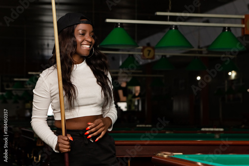 Young female billiard player holding a cue stick while standing next to a pool table with white balls in a dimly lit billiard room, creating a captivating atmosphere