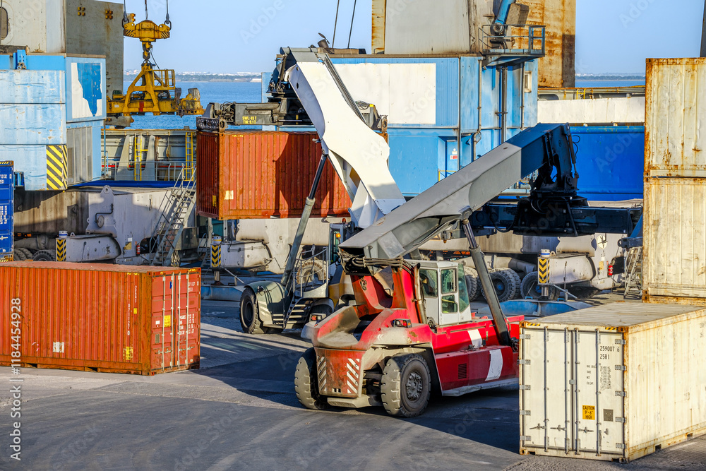 Reach stackers and cranes move containers near bulk carrier on sunny