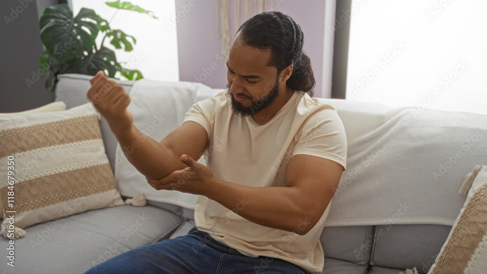 Man sitting on sofa indoors checks elbow for pain in modern living room with cushions around him showcasing cozy home interior ambiance. Man sitting on sofa indoors checks elbow for pain in modern living room with cushions around him showcasing cozy home interior ambiance.