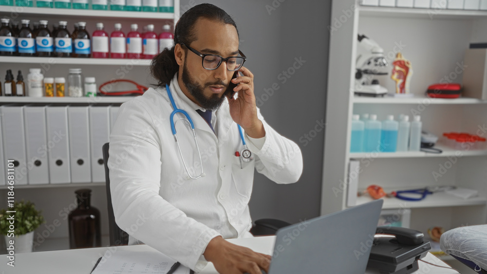 Man in clinic wearing glasses and lab coat talks on phone using laptop at desk surrounded by medical equipment and supplies in modern hospital interior. Man in clinic wearing glasses and lab coat talks on phone using laptop at desk surrounded by medical equipment and supplies in modern hospital interior.