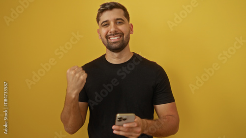 Young hispanic man smiling confidently holding phone against vibrant yellow background conveying joy and satisfaction photo