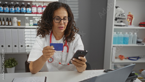 Woman in clinic holding credit card and smartphone wears glasses and stethoscope surrounded by medical supplies and white uniform focused on online transaction work
