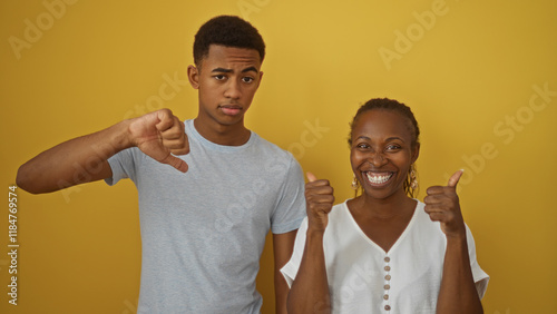 Mother and son making opposing thumb gestures in front of a yellow wall, expressing differing opinions with mother smiling and son serious photo