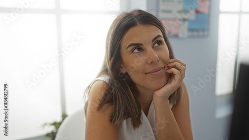 Free Download Young woman with brunette hair sitting in an office room, looking thoughtful and attractive, with a soft smile in an indoor workplace setting - GFXLoad