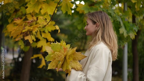 A cheerful young woman smiles widely while holding up vibrant and colorful autumn leaves in a beautiful fall setting