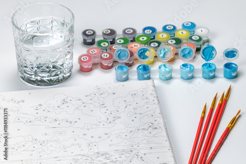 Painting by numbers kit featuring paint pots, brushes, canvas, and a glass of water on a white background. photo