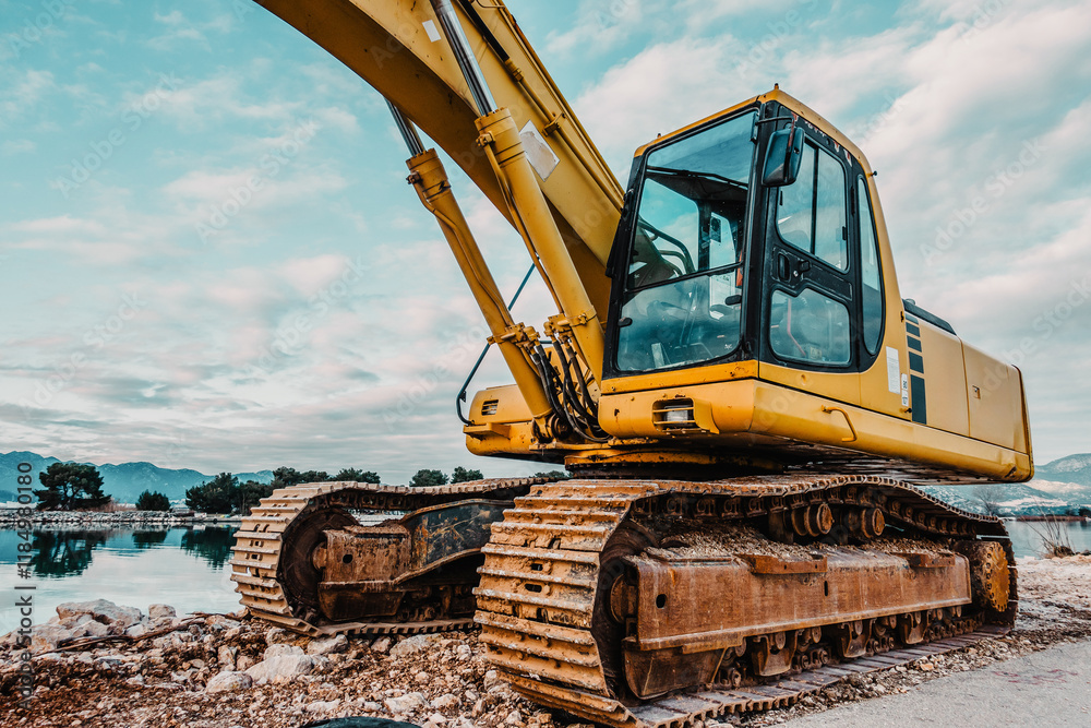 Fototapeta premium Heavy Yellow Excavator on Rocky Terrain at a Coastal Construction Site with Calm Water