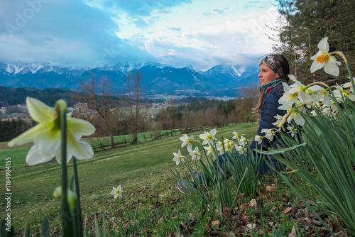 Hiker woman sitting in field of blooming daffodils with scenic view of snow capped mountain peaks of Karawanks seen from Kathreinkogel, Schiefling am See, Carinthia, Austria. Springtime Austrian Alps photo