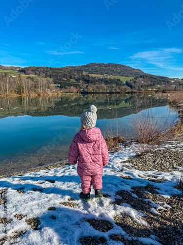 Cheerful toodler at shore of river Drava winding through Rosental valley. Snow-capped mountains of Karawanks, Sankt Jakob im Rosental, Carinthia, Austria. Winter wonderland in Austrian Alps. Childhood photo