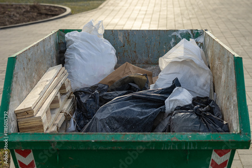 Close-up of a green industrial dumpster filled with construction debris, including wooden planks, black and white plastic garbage bags, and paper waste, placed outdoors