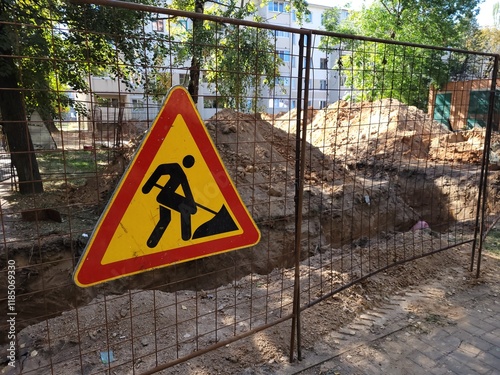 Construction site on city street is marked by metal barricade with triangular warning sign depicting road work